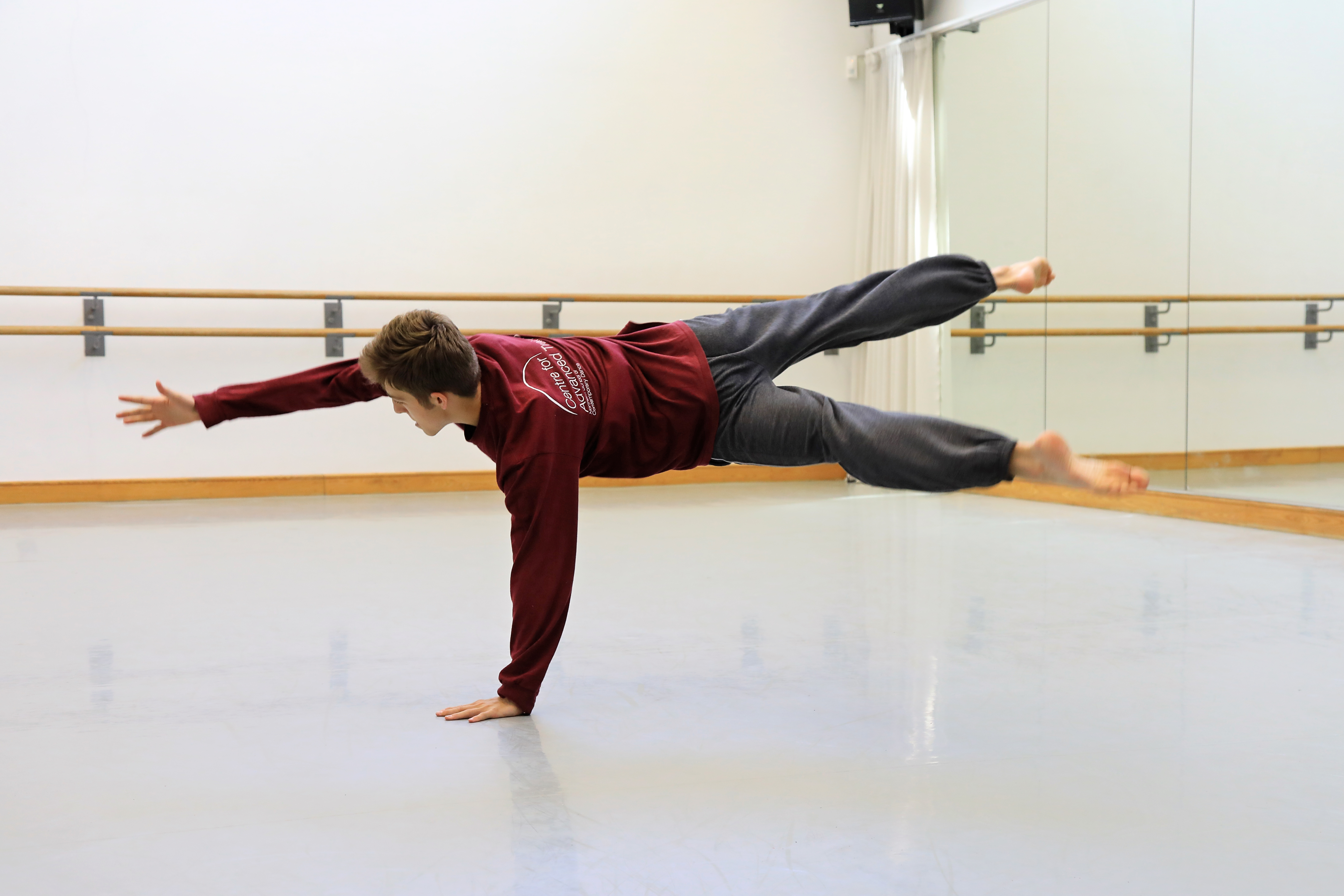 A dancer is in a studio with a grey floor, ballet barres and mirrors. They are in a press up position with one arm and both legs off the floor, there is a blur of their legs which shows the movement. 