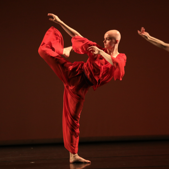 A dancer stands on a stage in a bright red costume. They are kicking one leg high up behind them with a bent knee and they are looking straight to the side. which twists their body towards the leg.