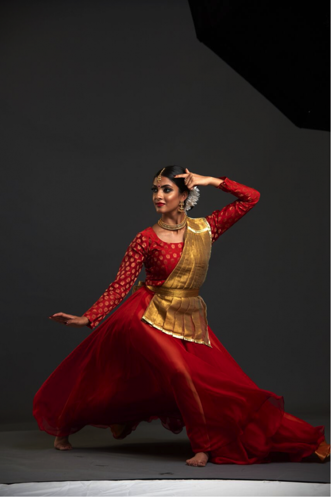 Vidya is performing on a black stage. They have their head turned to the left, with one finger touching the side of the head. They are wearing gold jewelry and red and gold clothing. Their legs are splayed wide under a large red skirt in movement.