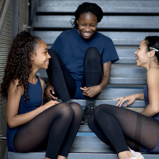 Three dancers are sat on a set of stairs, laughing and smiling at each other. They are wearing black tights and navy leotards and tops.