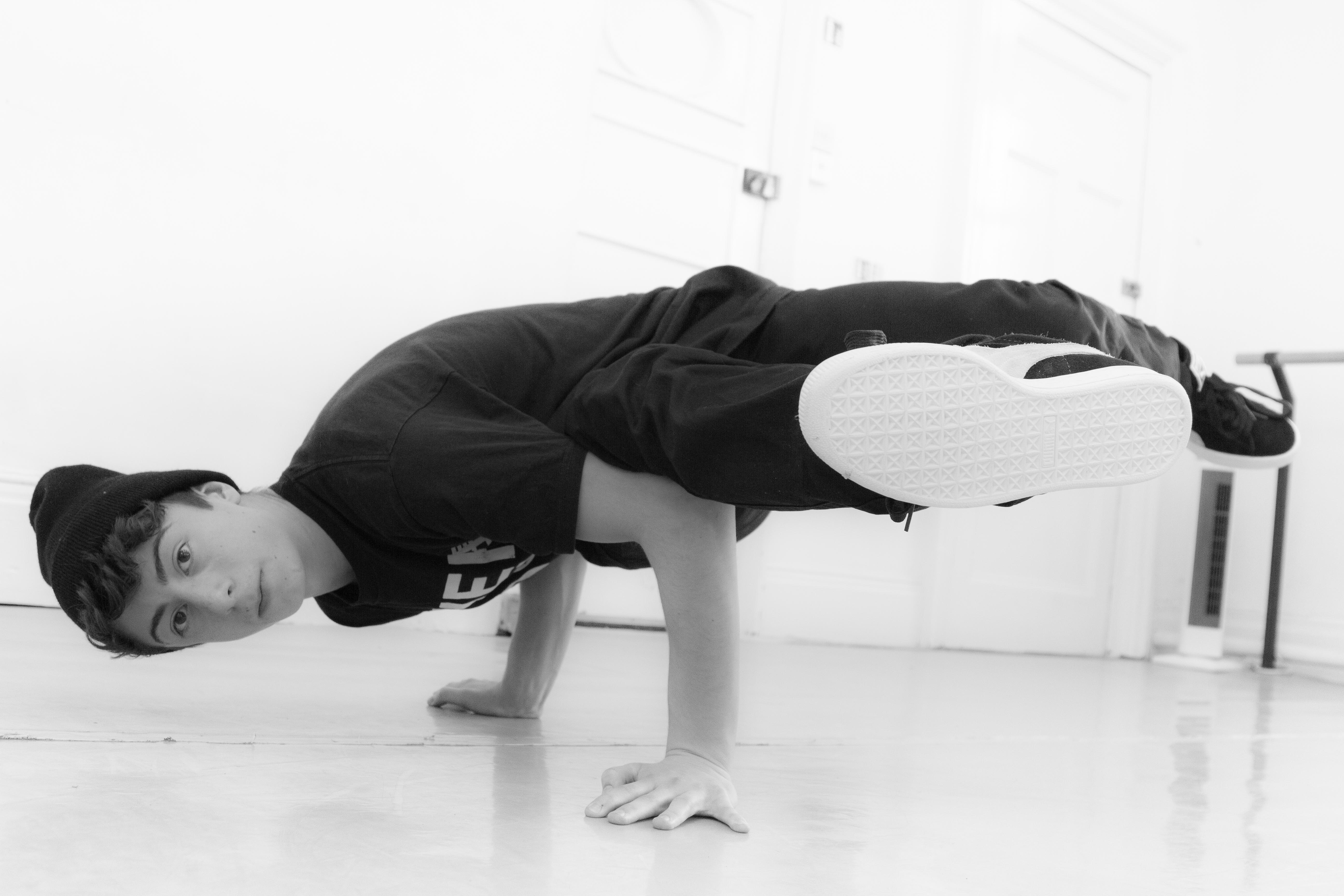 A black and white image of a break dancer, they are balanced on their hands with their legs bent up off the floor. They are wearing a beanie hat. 