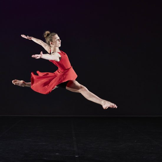 A dancer leaps high in the air with their arms thrown sideways. They are wearing a floaty red dress in contrast to the black background.