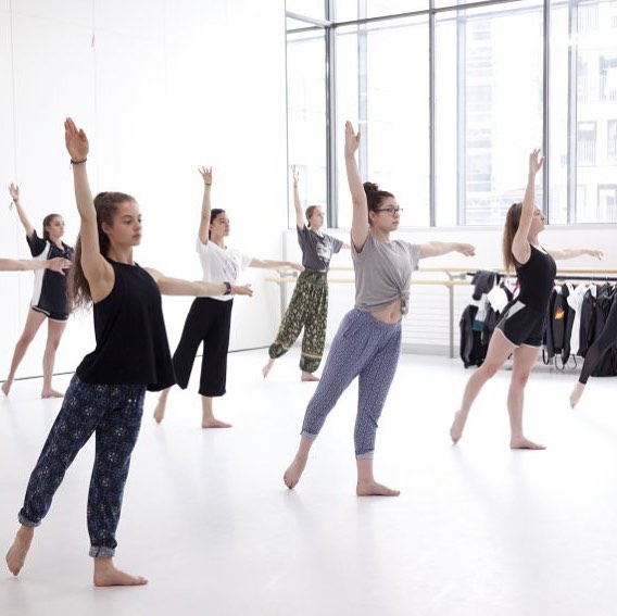 Six dancers are in the same position, they have one arm pointing towards the ceiling and one leg stretched behind them, they look strong and focused. There are large windows to the side and the studio is light.