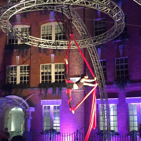 Photo by Liam Croucher Willow is hanging in the splits from red silks that are attached to a high metal rig. They are performing at night time outdoors. Behind them are large buildings dramatically lit by purple light.