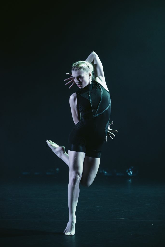 Hayley, a dancer in a black leotard is standing on a black stage. Blue light is shining on them. They are in a striking poised position. They are stood on one raised leg, the other is bent behind their front leg and is pointed sharply upwards. One arm is bent behind their back and their fingers are splayed outwards. The other arm is raised overhead and bent behind their head, with their fingers splayed outwards.