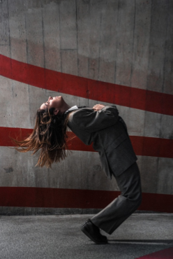 Annie is wearing a grey oversized suit. They are balanced with their back bent backwards and their heels just raised of the ground. They are against a grey concrete backdrop with red painted stripes.