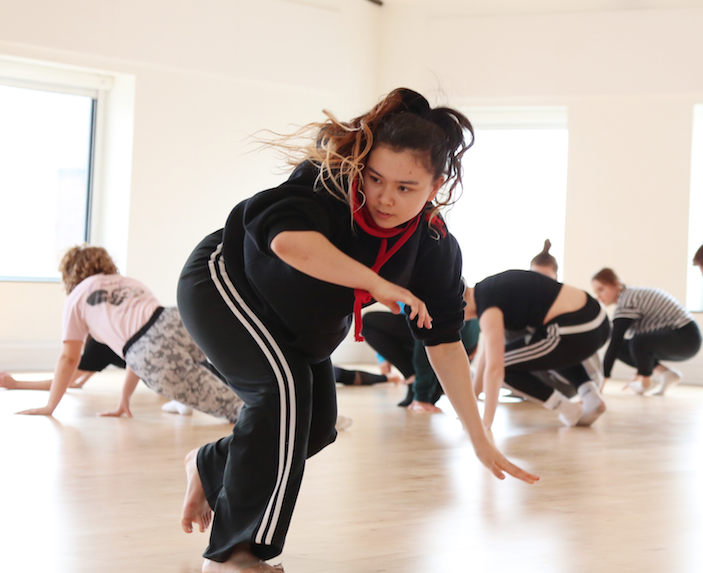 Sua is working in a studio. They are in the middle of a dynamic movement, about to crouch into the floor. They have a concentrated expression on their face. They are wearing tracksuit bottoms and a black and red hoodie.