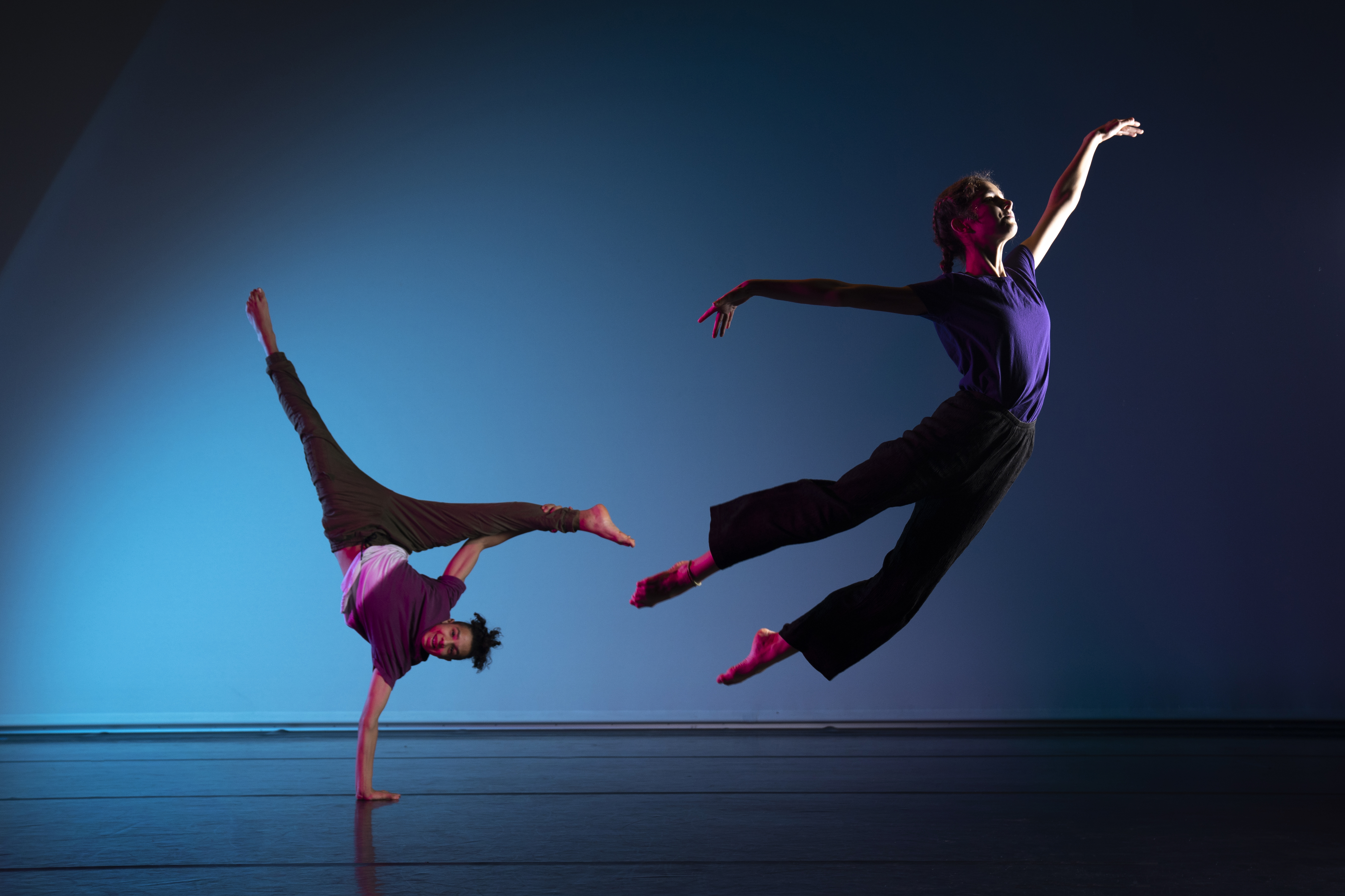 An image from a photoshoot. The background is blue and two dancers are positioned in front. One is balancing on one hand, and the other is gliding through the air. The lighting is dark and dramatic.