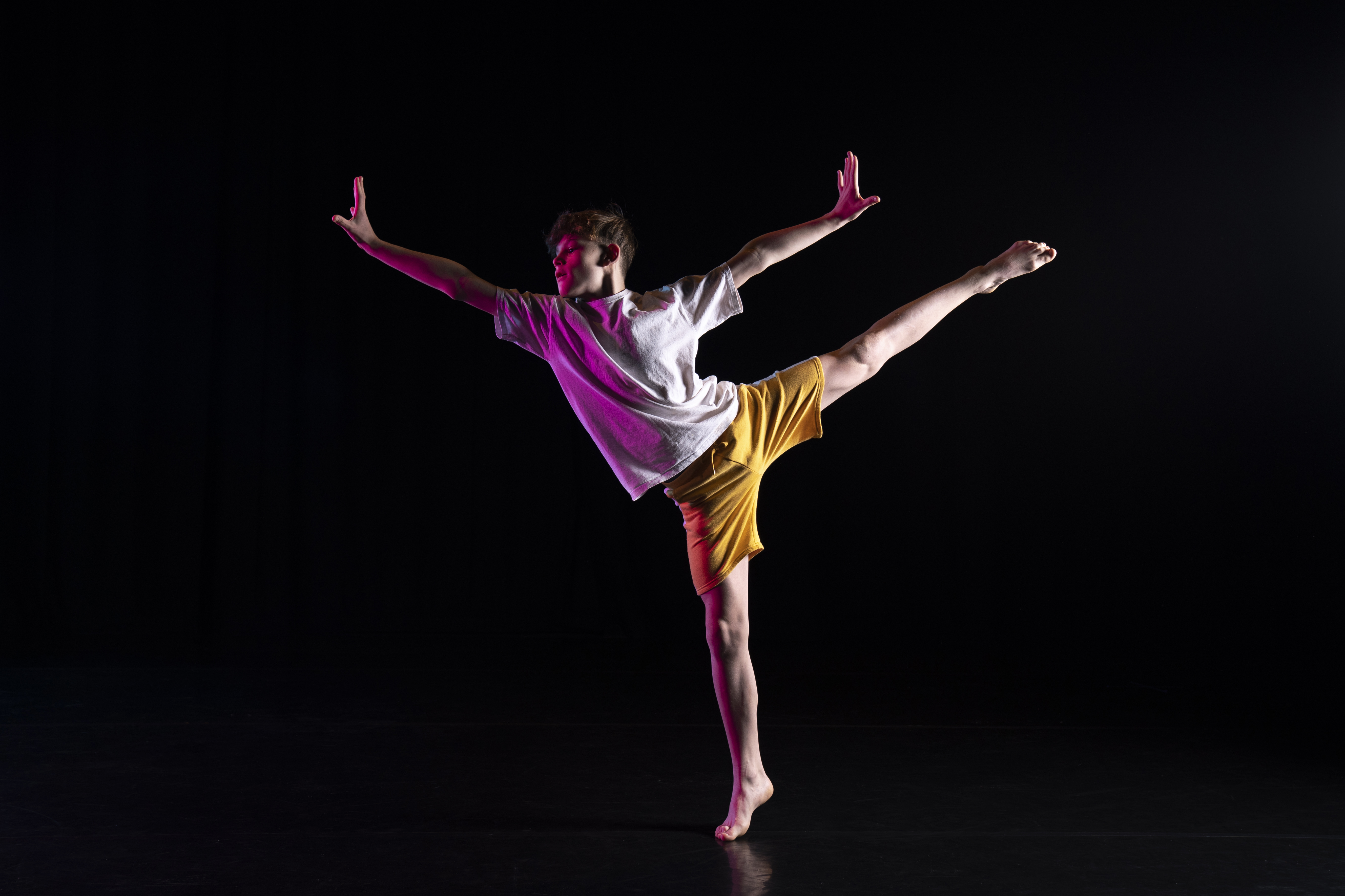 An image from a photoshoot. One male student is balancing on one leg whist reaching all his limbs away from his body. He is again a black background.