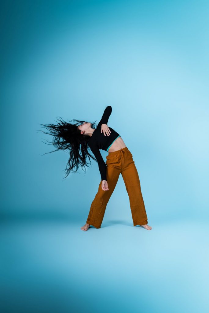 One female dancer leaning sideways with her elbow reaching in the air, her hair is swinging outwards. The background is bright blue